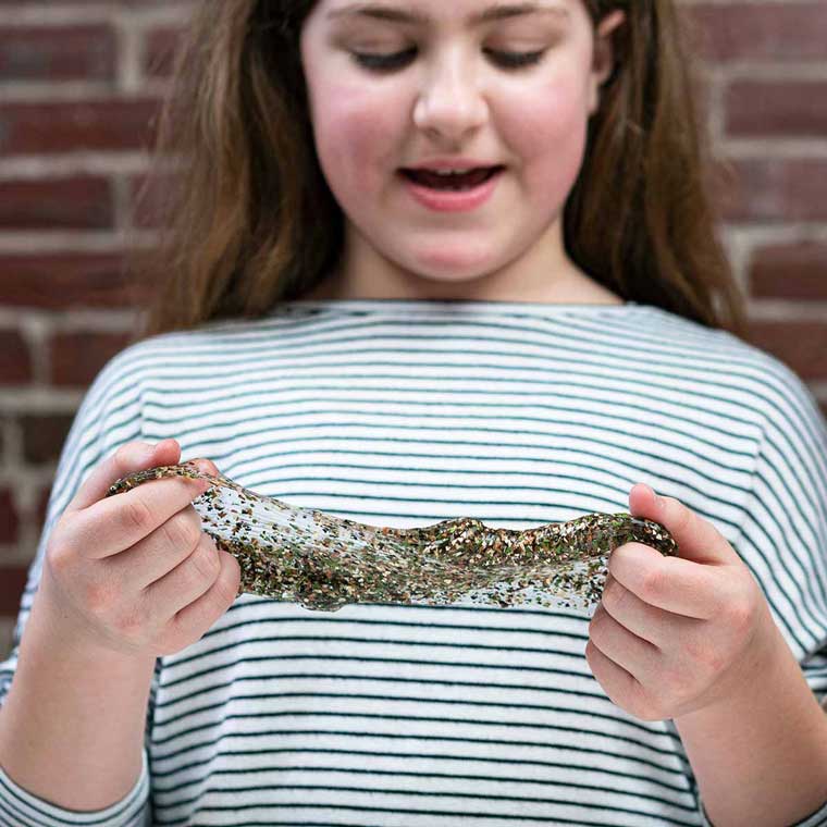 Girl standing near a brick background holding clear putty with camo sparkles.