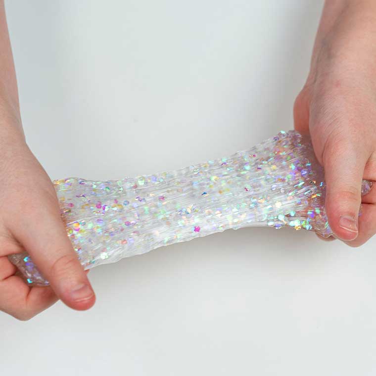 Two hands resting on a white surface stretching clear putty with rainbow glitter in it.